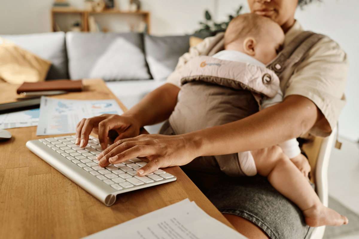 Young woman typing on keyboard while her baby sleeping in sling, she working at home during maternity leave
