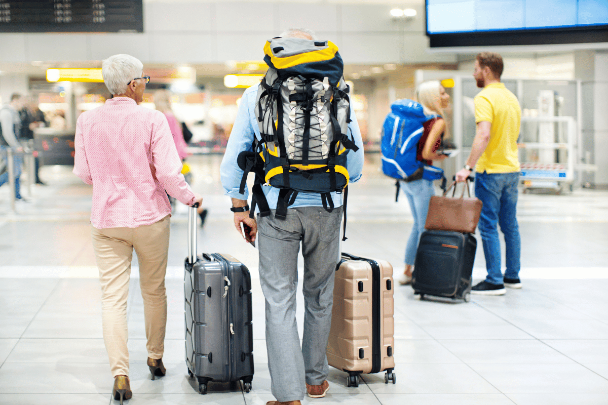 Closeup rear view of a senior couplepulling their suitcases and backpackstowards check in counter. There's ayoung couple in front of them waitingfor their flight.
